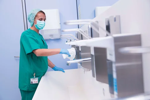 Eine Bild von einer Frau im grünen OP Kleidung mit Mundschutz die Seifen- und Desinfektionsspender wischt