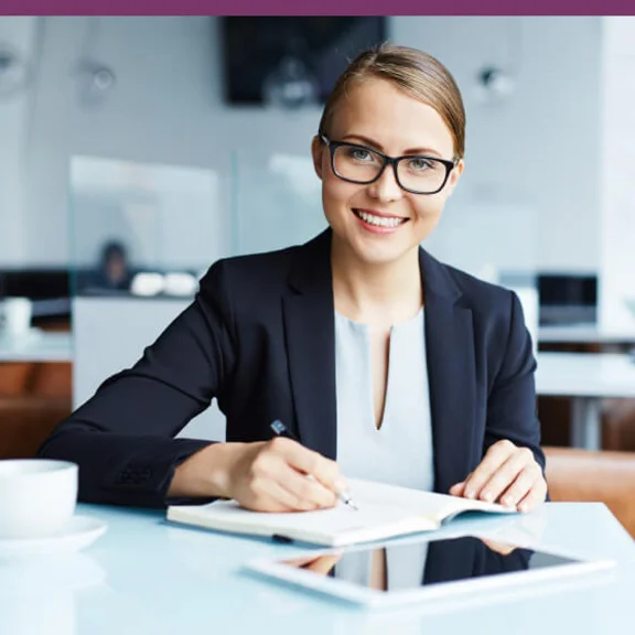 Eine weibliche Person sitzt schreibend an einem Schreibtisch und lächelt.