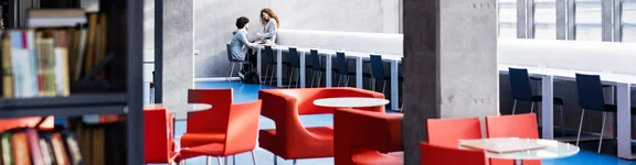 Ein Bild in einer Bibliothek, mit Bücherregalen und kleinen Sitzecken mit roten Sesseln, im Hintergrund ist ein Mann und eine Frau die sich gegenüber sitzen