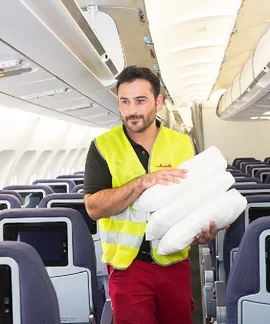 Klüh Airport-Service Angestellter in Arbeitskleidung mit roter Hose und Gelber Warnweste mit Klüh Logo in einem Flugzeug mit 3 weißen Kopfkissen in seiner Hand